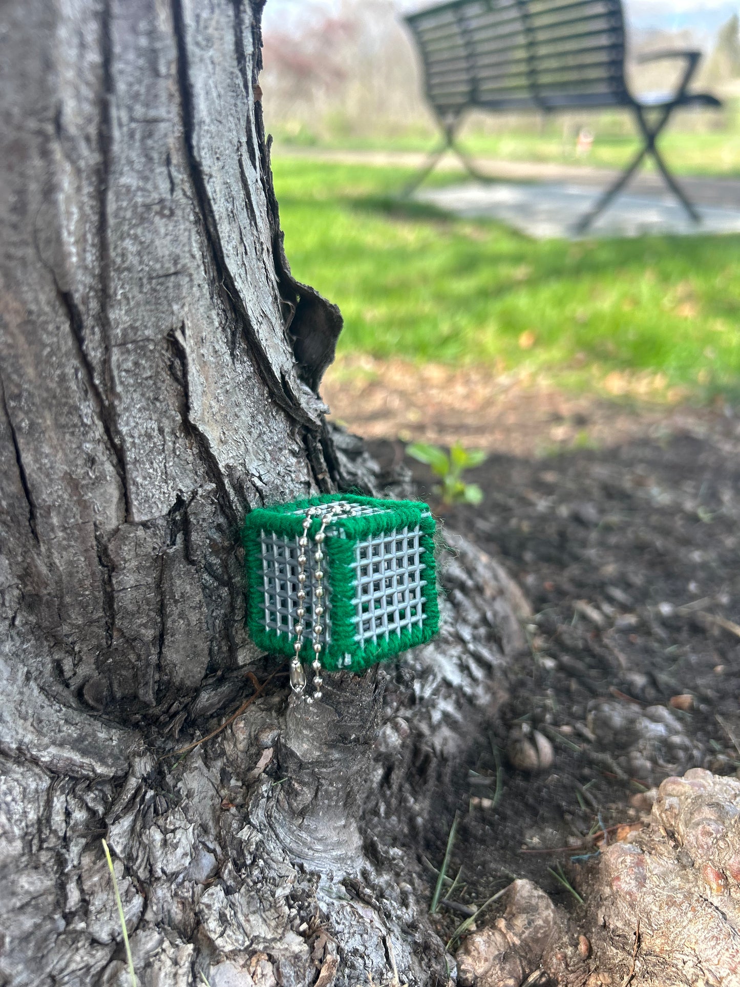 Handmade ASMR sensory cube laced with green yarn, 1.25 in. hexagon fidget art with ball chain clip pictured at the base of a tree on one of the raised toots behind a black metal park bench.