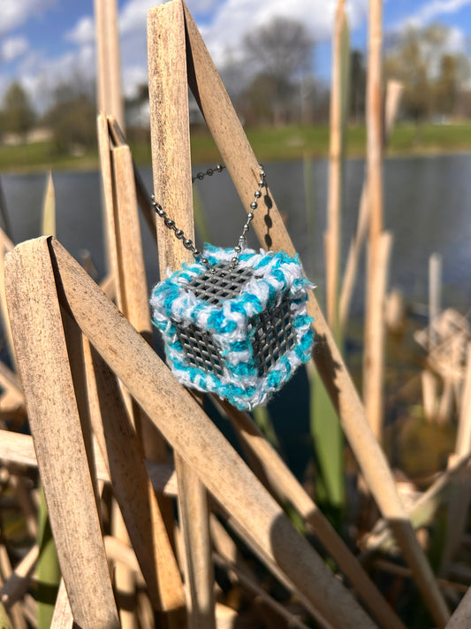 Handmade ASMR sensory cube laced with soft blue and white yarn, 1.25 in. hexagon fidget art with ball chain clip pictured in the reeds by a lake.