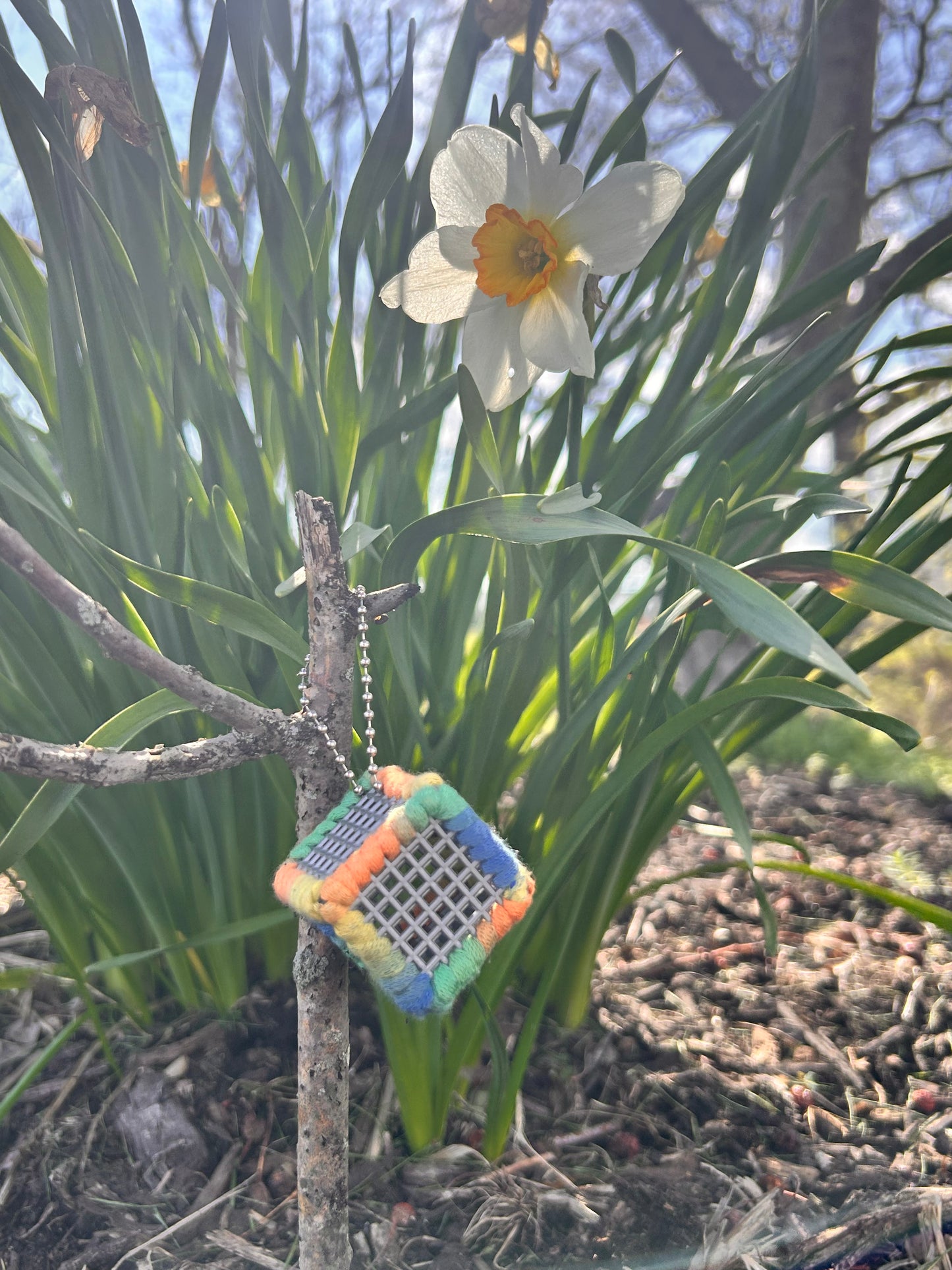 Handmade ASMR sensory cube laced with rainbow-inspired yarn, 1.25 in. hexagon fidget art with ball chain clip hanging from a stick in front of a white and yellow daffodil on a sunny day.