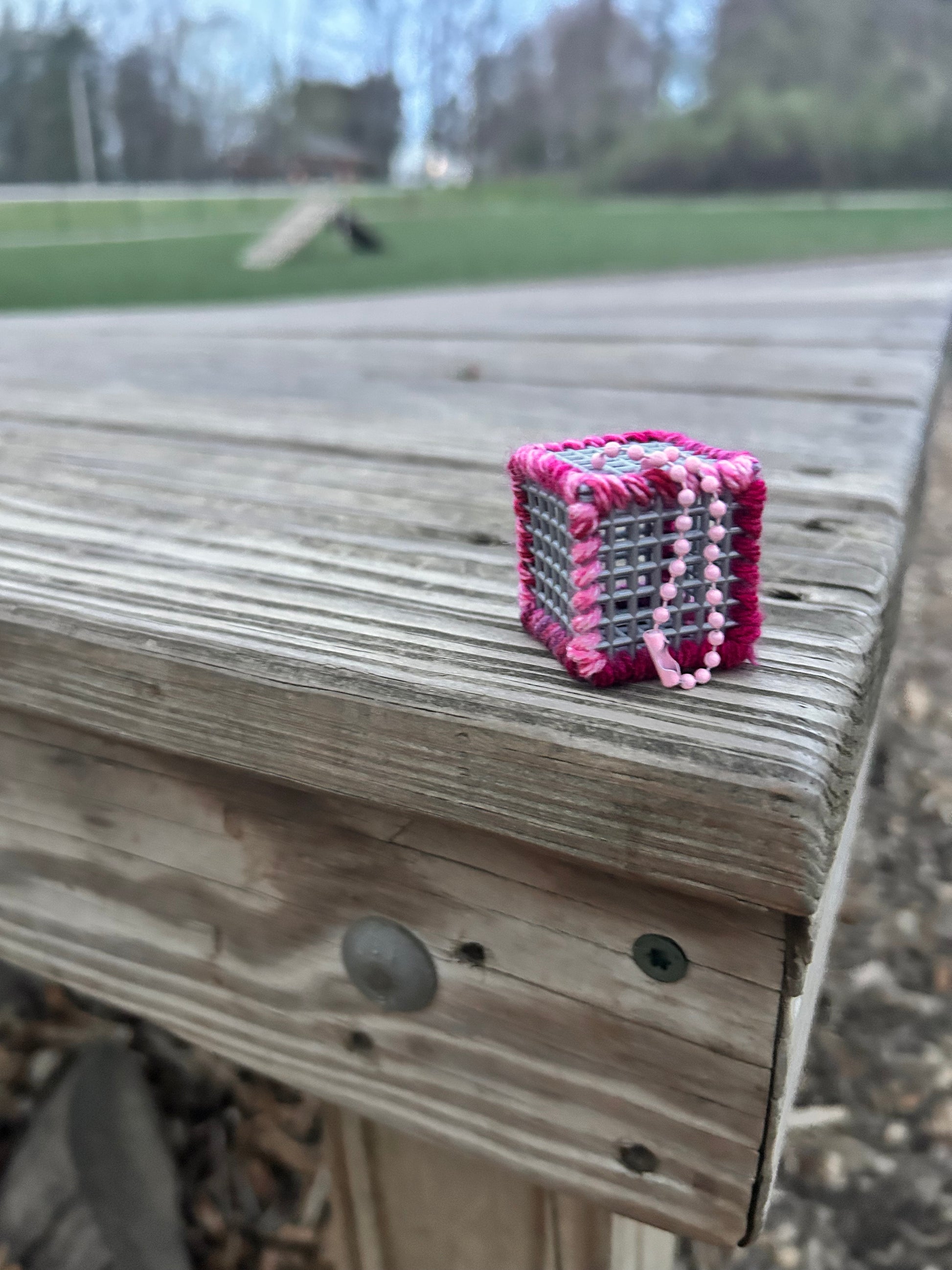 Handmade ASMR sensory cube laced with varied shades of pink and purple yarn, 1.25 in. hexagon fidget art with ball chain clip pictured on a wooden platform at the dog park.
