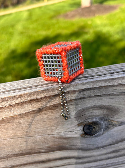 Handmade ASMR sensory cube laced with orange glitter yarn, 1.25 in. hexagon fidget art with ball chain clip pictured on a wooden fence slat with lush sunny green grass in the background.