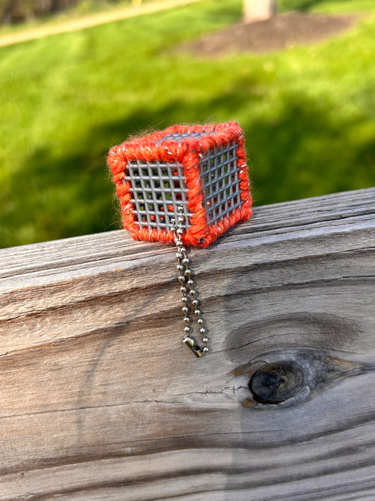 Handmade ASMR sensory cube laced with orange glitter yarn, 1.25 in. hexagon fidget art with ball chain clip pictured on a wooden fence slat with lush sunny green grass in the background.