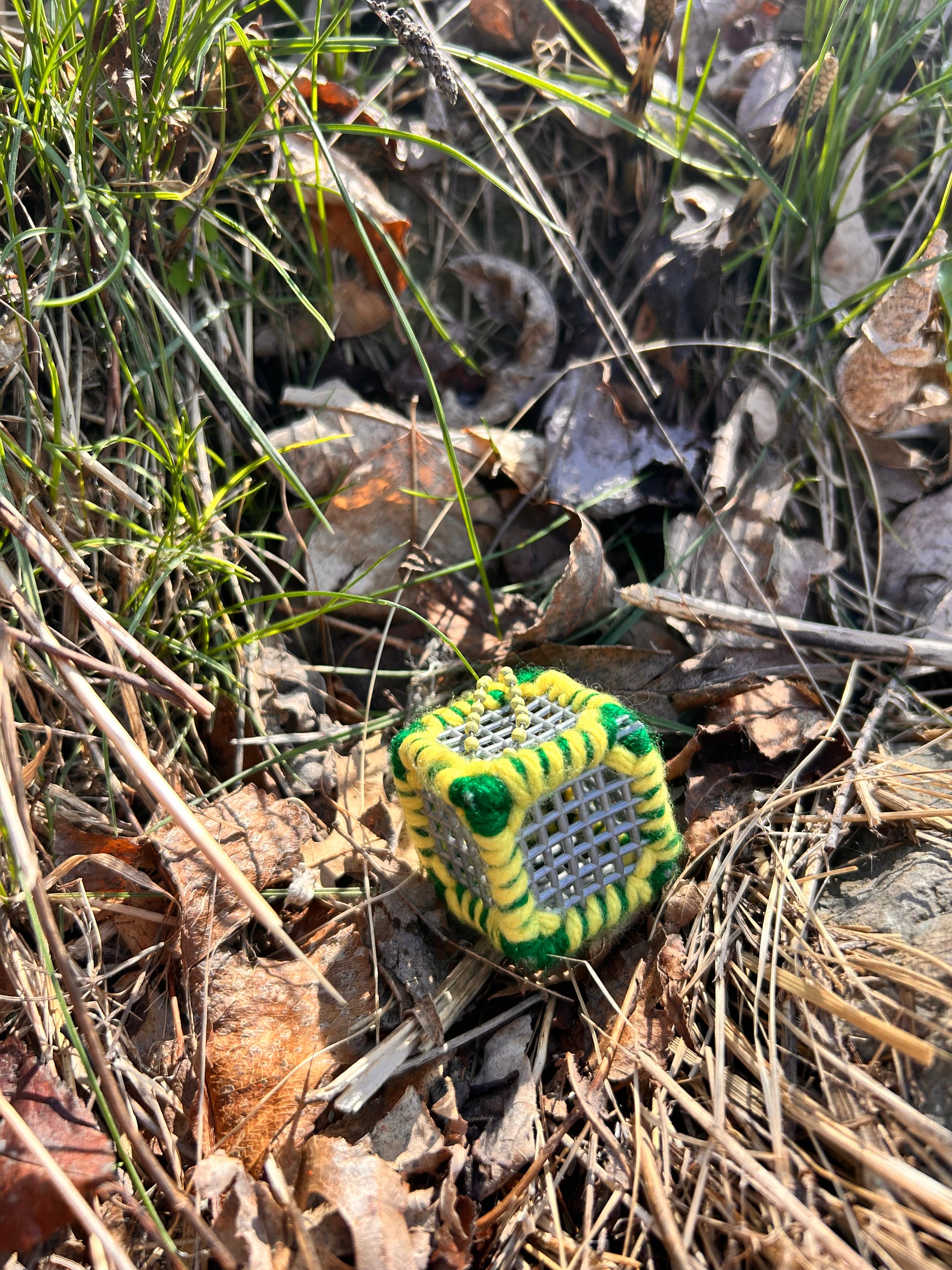Handmade ASMR sensory cube laced with green and yellow yarn, 1.25 in. hexagon fidget art with ball chain clip pictured in the grass on a sunny day with some dried-up leaves at the edge of a wooded area.