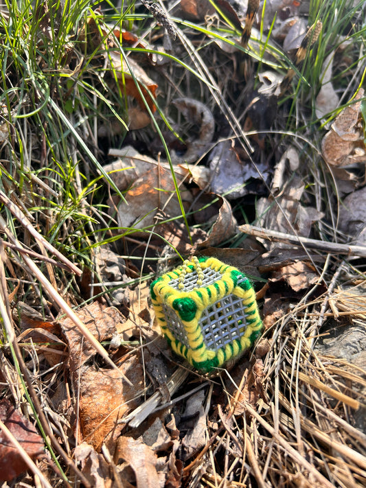 Handmade ASMR sensory cube laced with green and yellow yarn, 1.25 in. hexagon fidget art with ball chain clip pictured in the grass on a sunny day with some dried-up leaves at the edge of a wooded area.