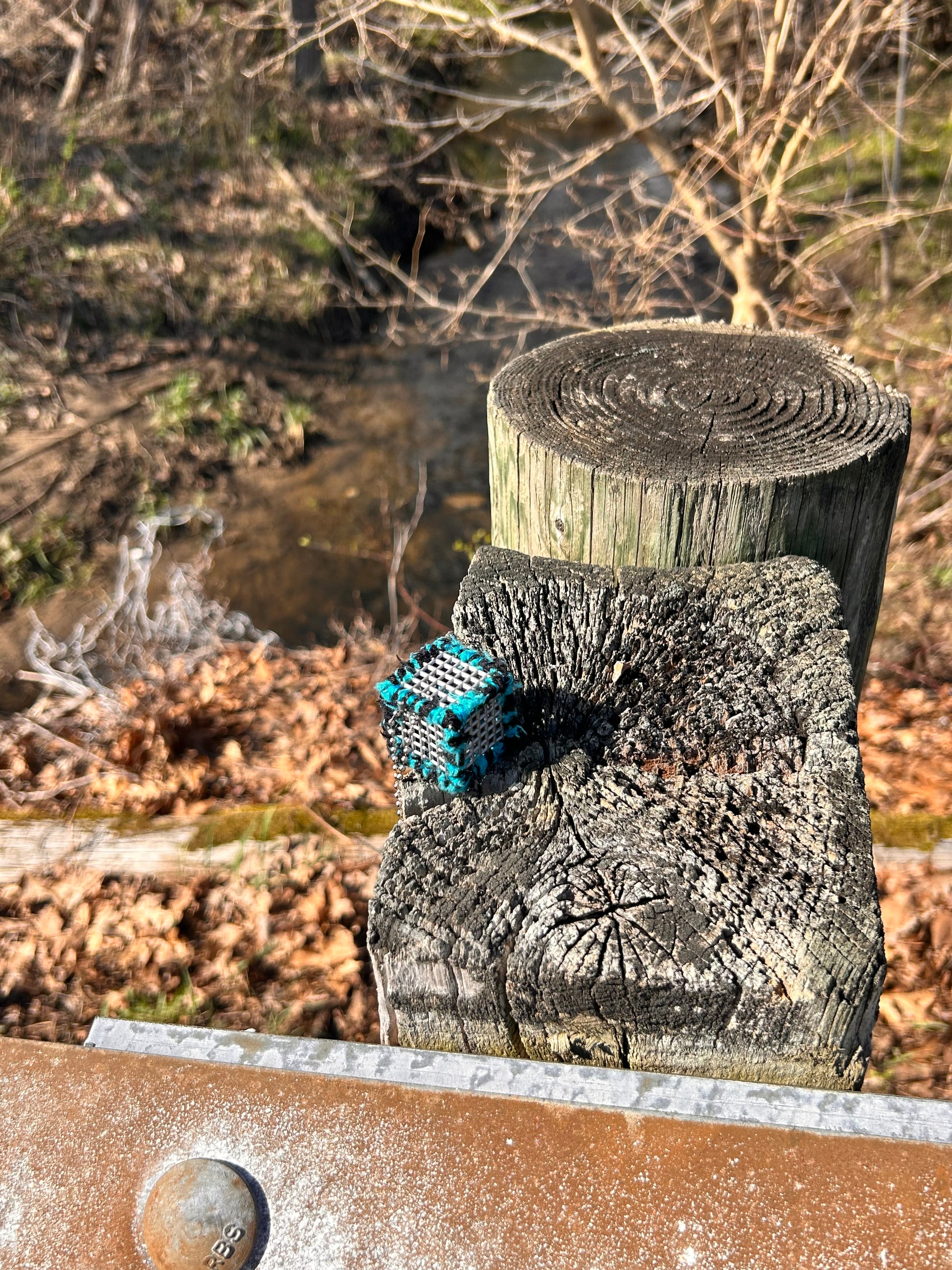 Handmade ASMR sensory cube laced with soft textured teal blue and black yarn, 1.25 in. hexagon fidget art with ball chain clip pictured on a wooden post near the edge of the road by a stream.