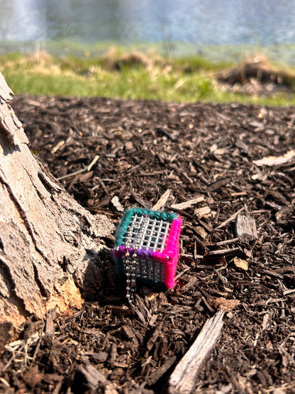 Handmade ASMR sensory cube laced with variegated green, purple and pink yarn, 1.25 in. hexagon fidget art with ball chain clip pictured at the base of a tree on brown mulch with a grassy area and lake in the background.
