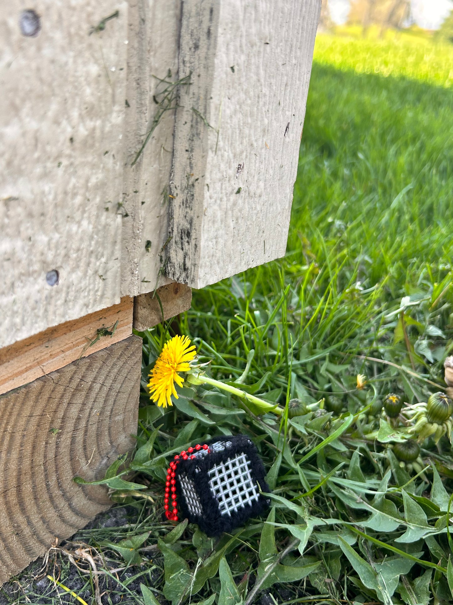 Handmade ASMR sensory cube laced with soft textured black yarn, 1.25 in. hexagon fidget art with ball chain clip pictured in the grass next to a dandelion bloom on the back side of a shed.