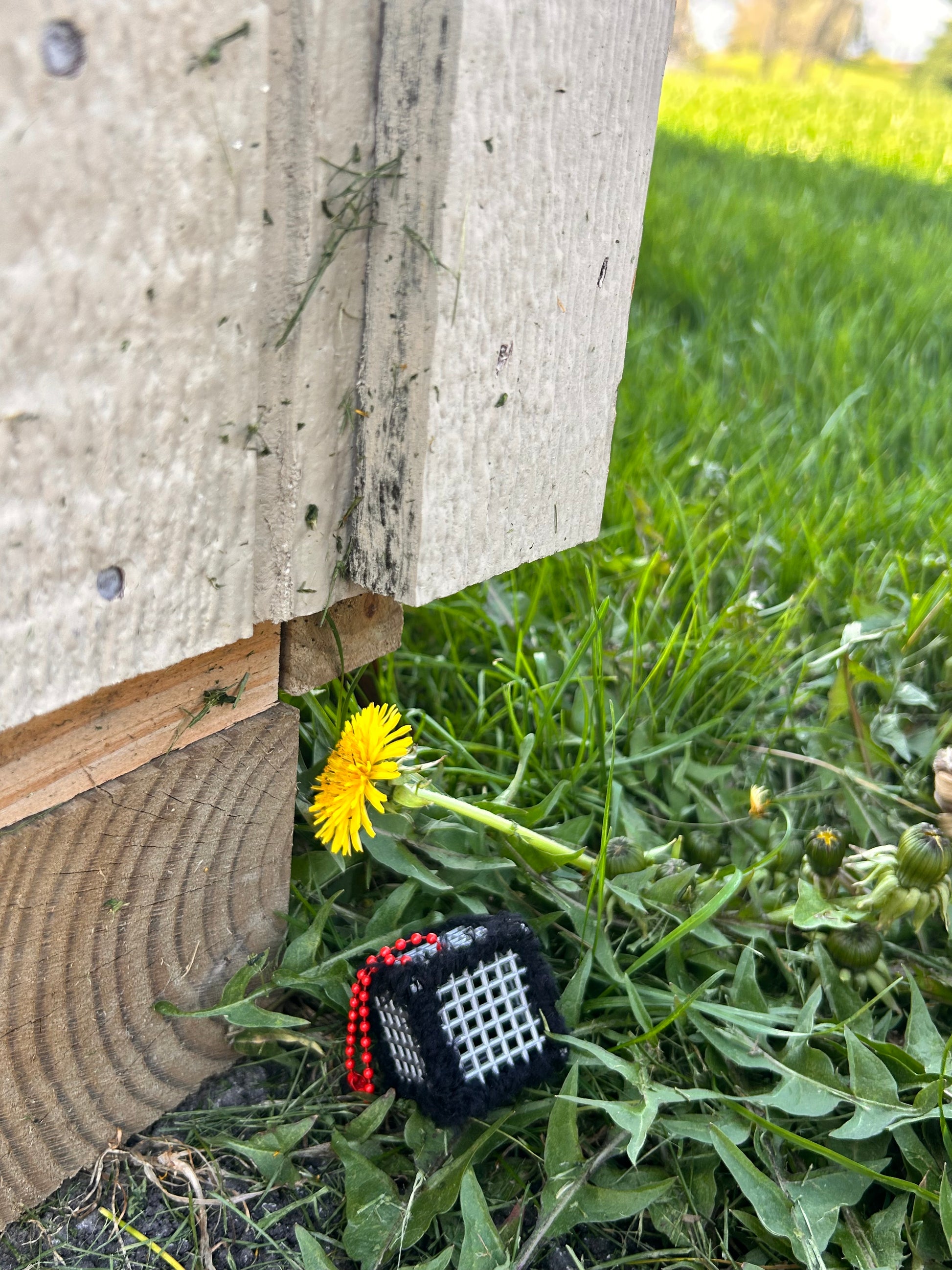 Handmade ASMR sensory cube laced with soft textured black yarn, 1.25 in. hexagon fidget art with ball chain clip pictured in the grass next to a dandelion bloom on the back side of a shed.