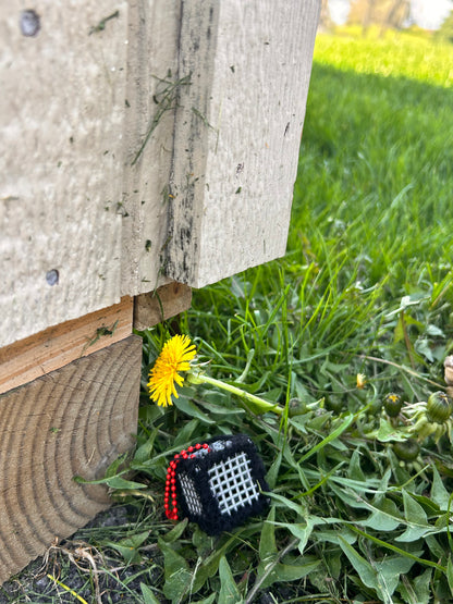 Handmade ASMR sensory cube laced with soft textured black yarn, 1.25 in. hexagon fidget art with ball chain clip pictured in the grass next to a dandelion bloom on the back side of a shed.