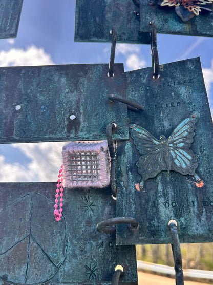 Handmade ASMR sensory cube laced with pink glitter yarn, 1.25 in. hexagon fidget art with ball chain clip pictured wedged in a gap of a blue and gray squared metal art collage with a metal butterfly beside it on one of the metal squares with beautiful blue skies and puffy white clouds in the background.