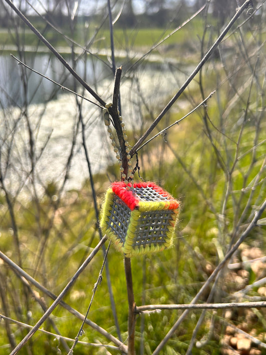 Handmade ASMR sensory cube laced with bright yellow and orange yarn, 1.25 in. hexagon fidget art with ball chain clip pictured on a tree branch near a lake.