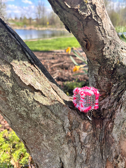 Handmade ASMR sensory cube laced with soft textured hot pink and white yarn, 1.25 in. hexagon fidget art with ball chain clip pictured in the fork of a tree on a sunny day with a flower bed of daffodils, green grass and a lake in the background .