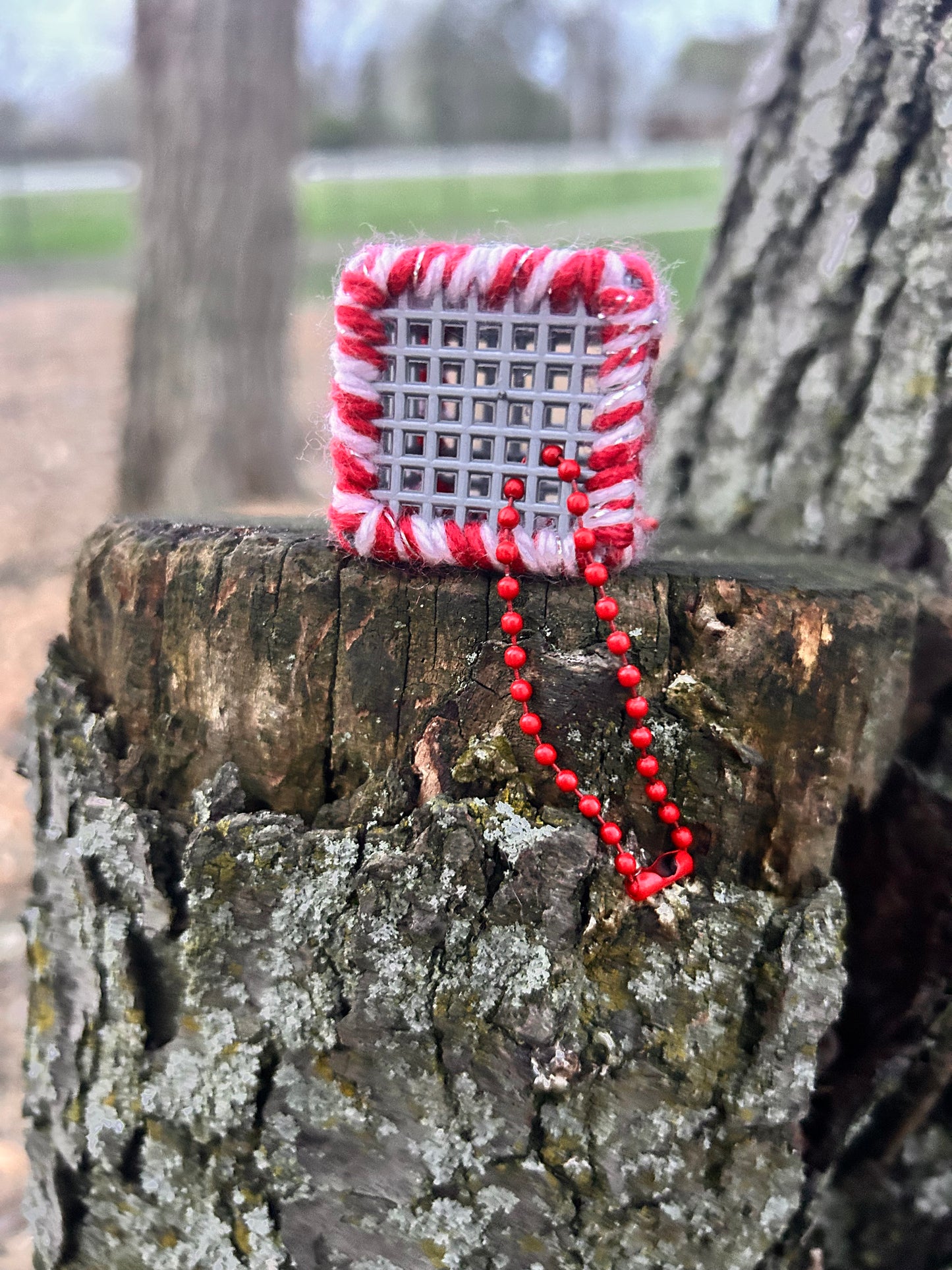 Handmade ASMR sensory cube laced with red and a pinky white glitter yarn, 1.25 in. hexagon fidget art with ball chain clip pictured on a tree stump in nature.
