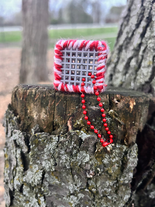 Handmade ASMR sensory cube laced with red and a pinky white glitter yarn, 1.25 in. hexagon fidget art with ball chain clip pictured on a tree stump in nature.