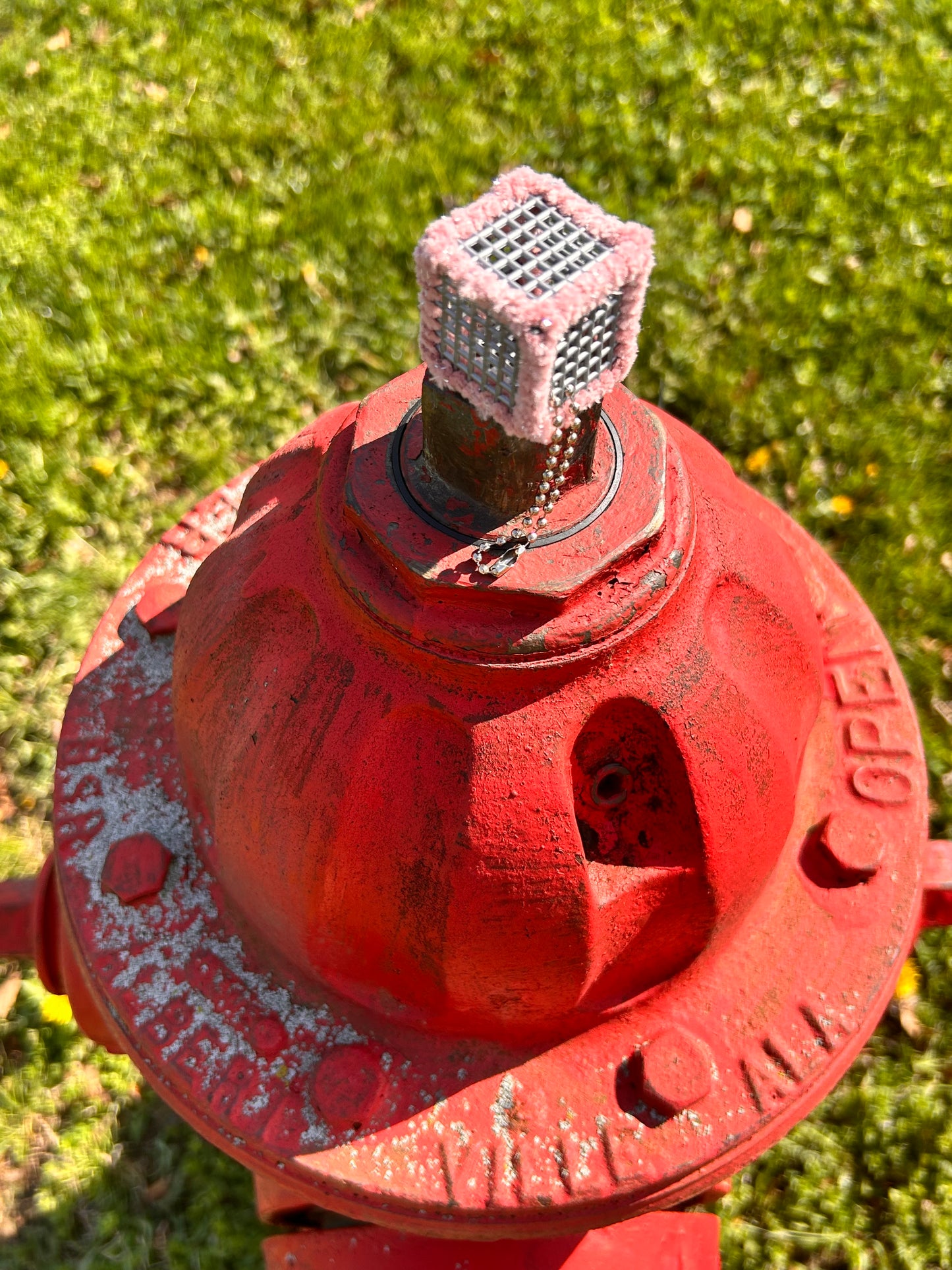 Handmade ASMR sensory cube laced with velvety soft pink glittery yarn, 1.25 in. hexagon fidget art with ball chain clip pictured atop a fire hydrant on a sunny day.