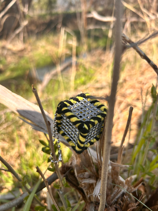 Handmade ASMR sensory cube laced with black and yellow yarn, 1.25 in. hexagon fidget art with ball chain clip nestled amongst the weeds with a stream running in the background.