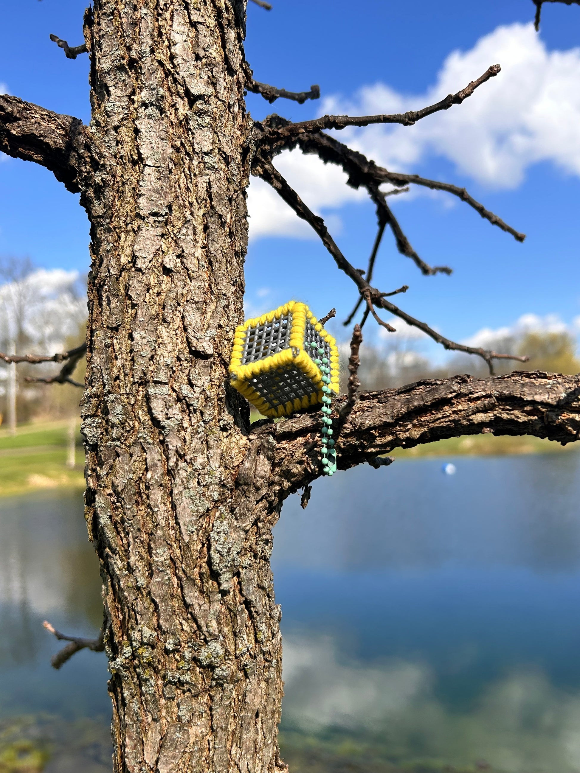 Handmade ASMR sensory cube laced with yellow yarn, 1.25 in. hexagon fidget art with ball chain clip pictured on the main branch of a tree on a sunny day with blue skies, fluffy white clouds, and a blurred background of trees around a lake.