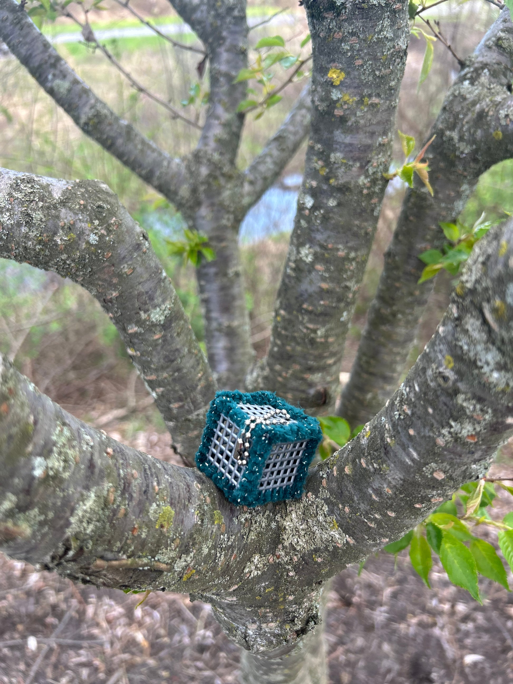 Handmade sensory ASMR cube wrapped in soft green with silver glitter yarn with ball chain clip nestled in the scaffold branches of a tree with a stream and reeds in the background.