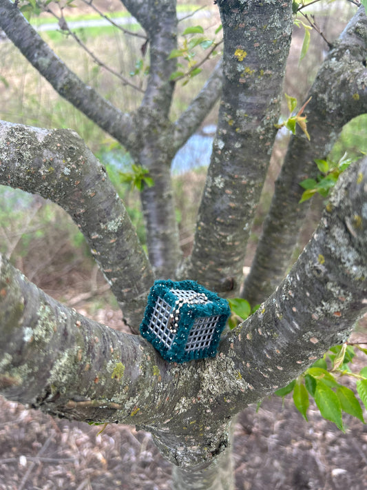Handmade sensory ASMR cube wrapped in soft green with silver glitter yarn with ball chain clip nestled in the scaffold branches of a tree with a stream and reeds in the background.