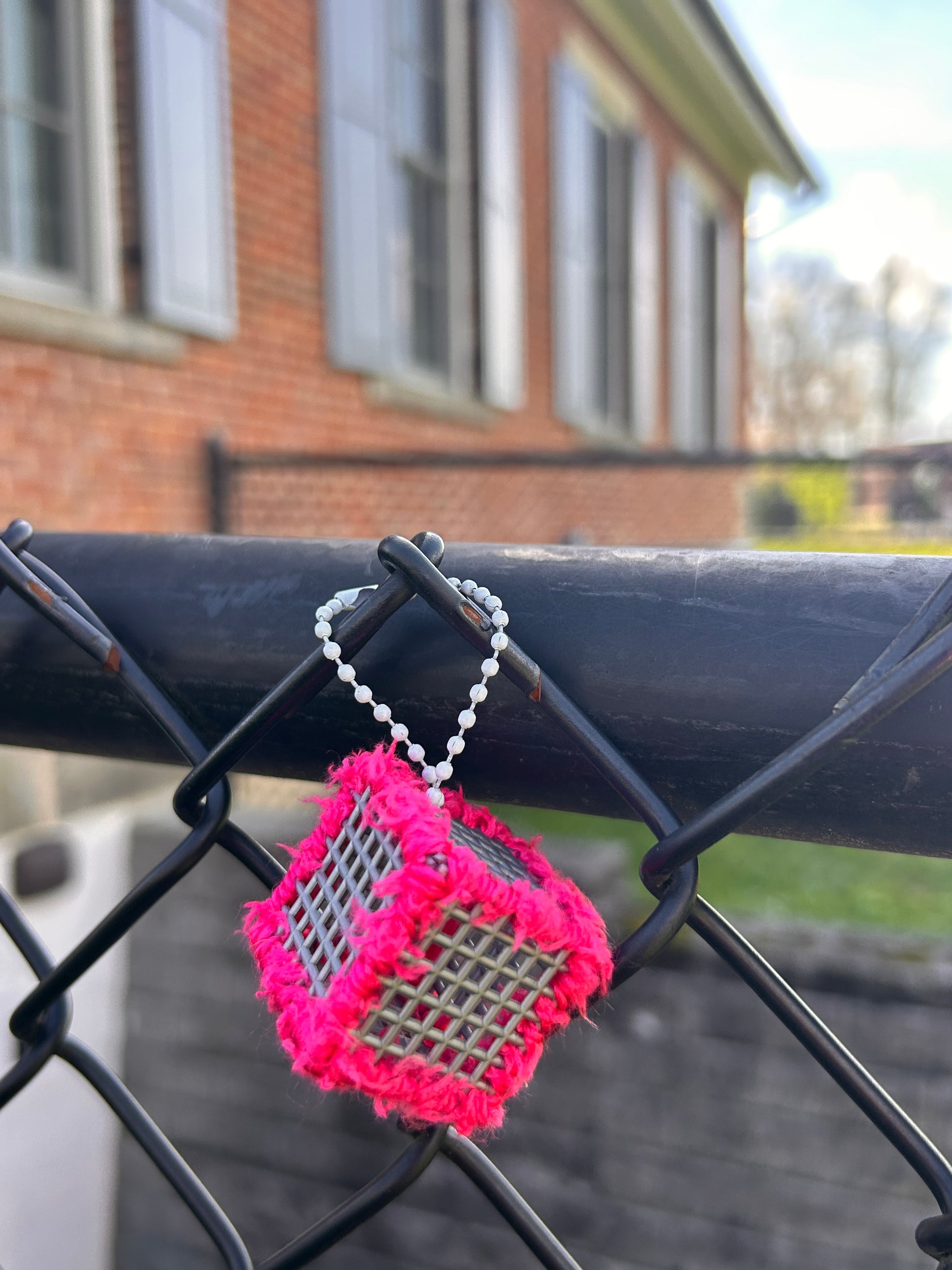 Handmade ASMR sensory cube laced with soft textured hot pink yarn, 1.25 in. hexagon fidget art with ball chain clip pictured hanging on a chain link fence outside a school building.
