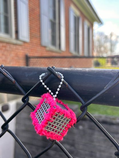 Handmade ASMR sensory cube laced with soft textured hot pink yarn, 1.25 in. hexagon fidget art with ball chain clip pictured hanging on a chain link fence outside a school building.