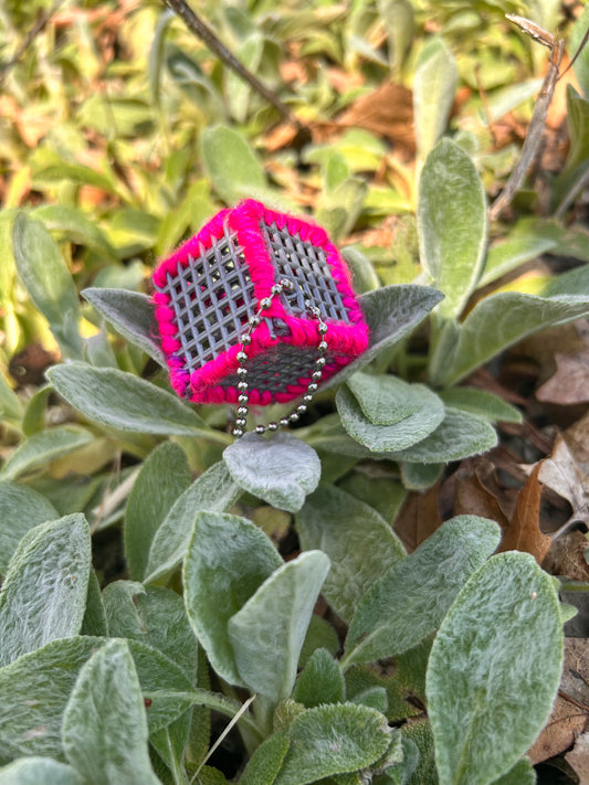 Handmade ASMR sensory cube laced with neon pink yarn, 1.25 in. hexagon fidget art with ball chain clip pictured sitting nestled softly on top of a patch of lamb's ear ground covering. 