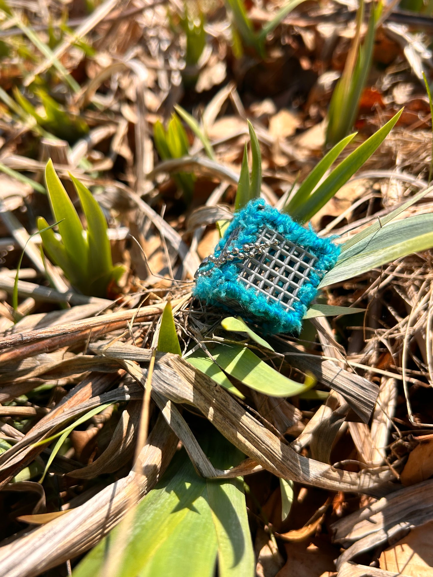 Handmade ASMR sensory cube laced with soft textured turquois blue yarn, 1.25 in. hexagon fidget art with ball chain clip nestles amongst newly emerged and dried out tiger lily leaves. 