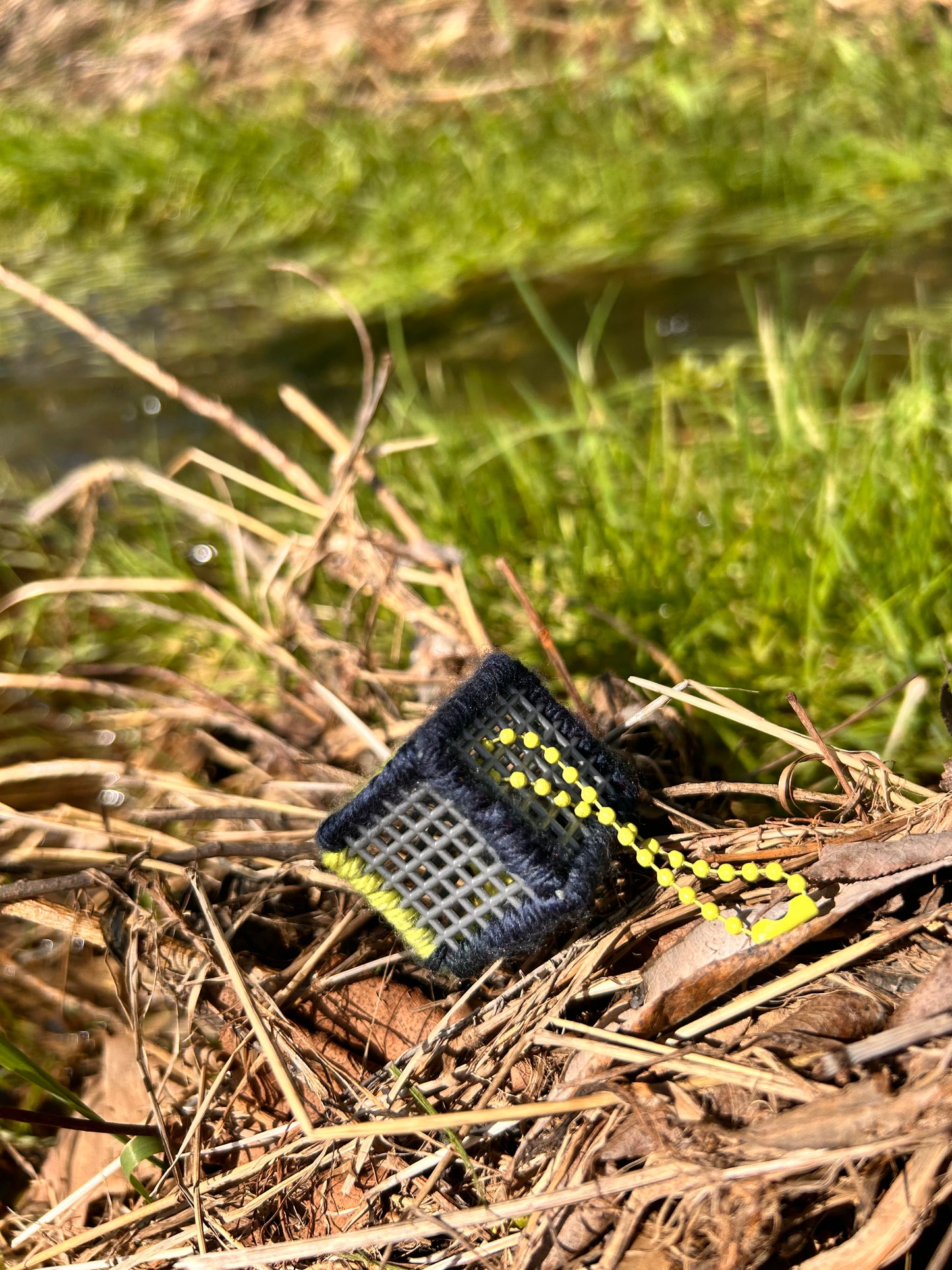 Handmade ASMR sensory cube laced with navy blue and yellow yarn, 1.25 in. hexagon fidget art with ball chain clip pictured sitting on a dried-up patch of weeds with lush green grass in the background.