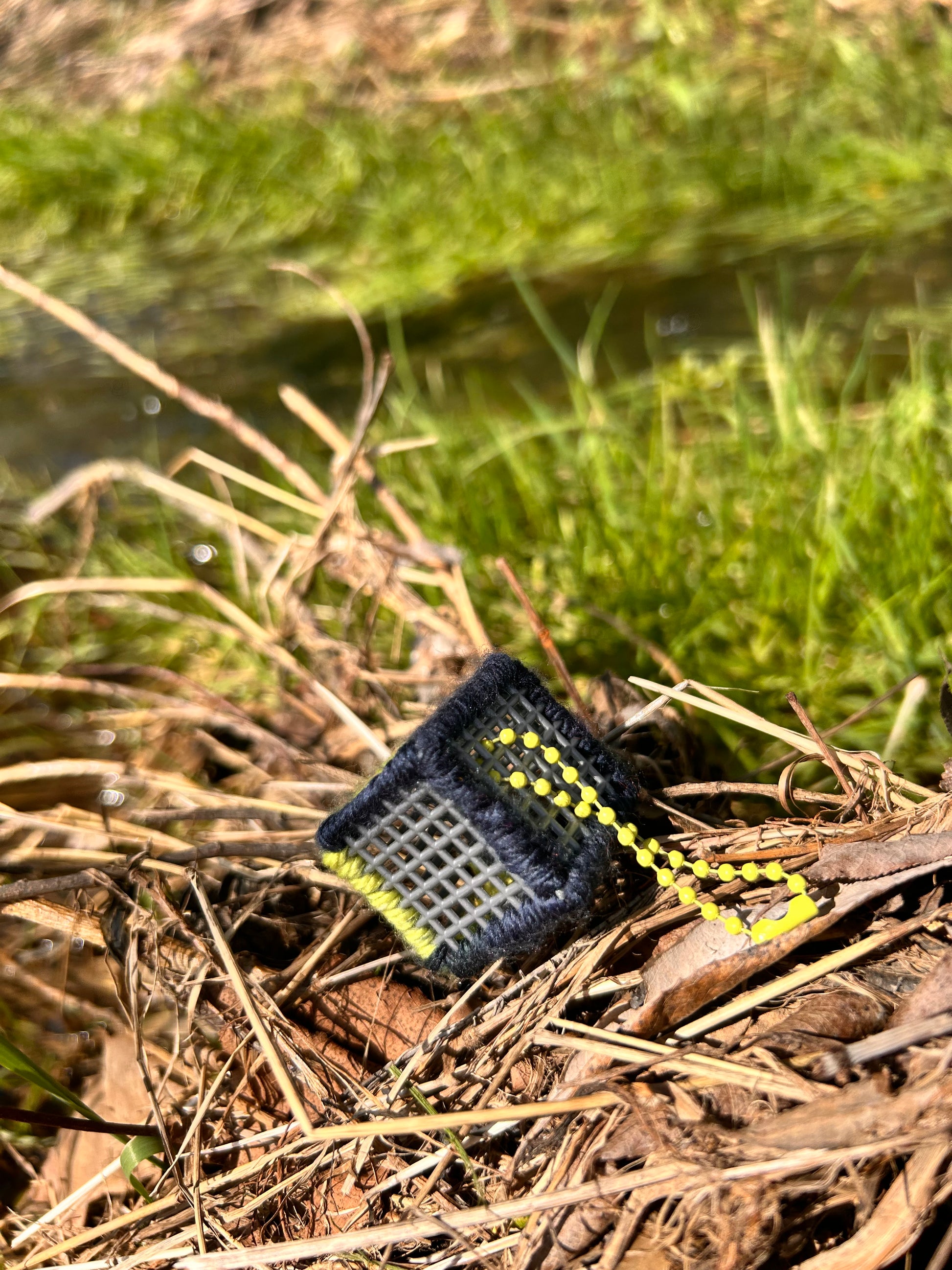 Handmade ASMR sensory cube laced with navy blue and yellow yarn, 1.25 in. hexagon fidget art with ball chain clip pictured sitting on a dried-up patch of weeds with lush green grass in the background.