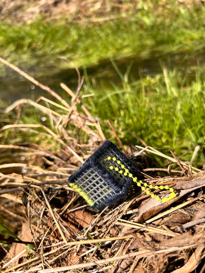 Handmade ASMR sensory cube laced with navy blue and yellow yarn, 1.25 in. hexagon fidget art with ball chain clip pictured sitting on a dried-up patch of weeds with lush green grass in the background.