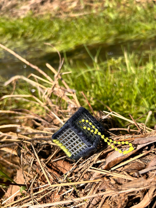 Handmade ASMR sensory cube laced with navy blue and yellow yarn, 1.25 in. hexagon fidget art with ball chain clip pictured sitting on a dried-up patch of weeds with lush green grass in the background.
