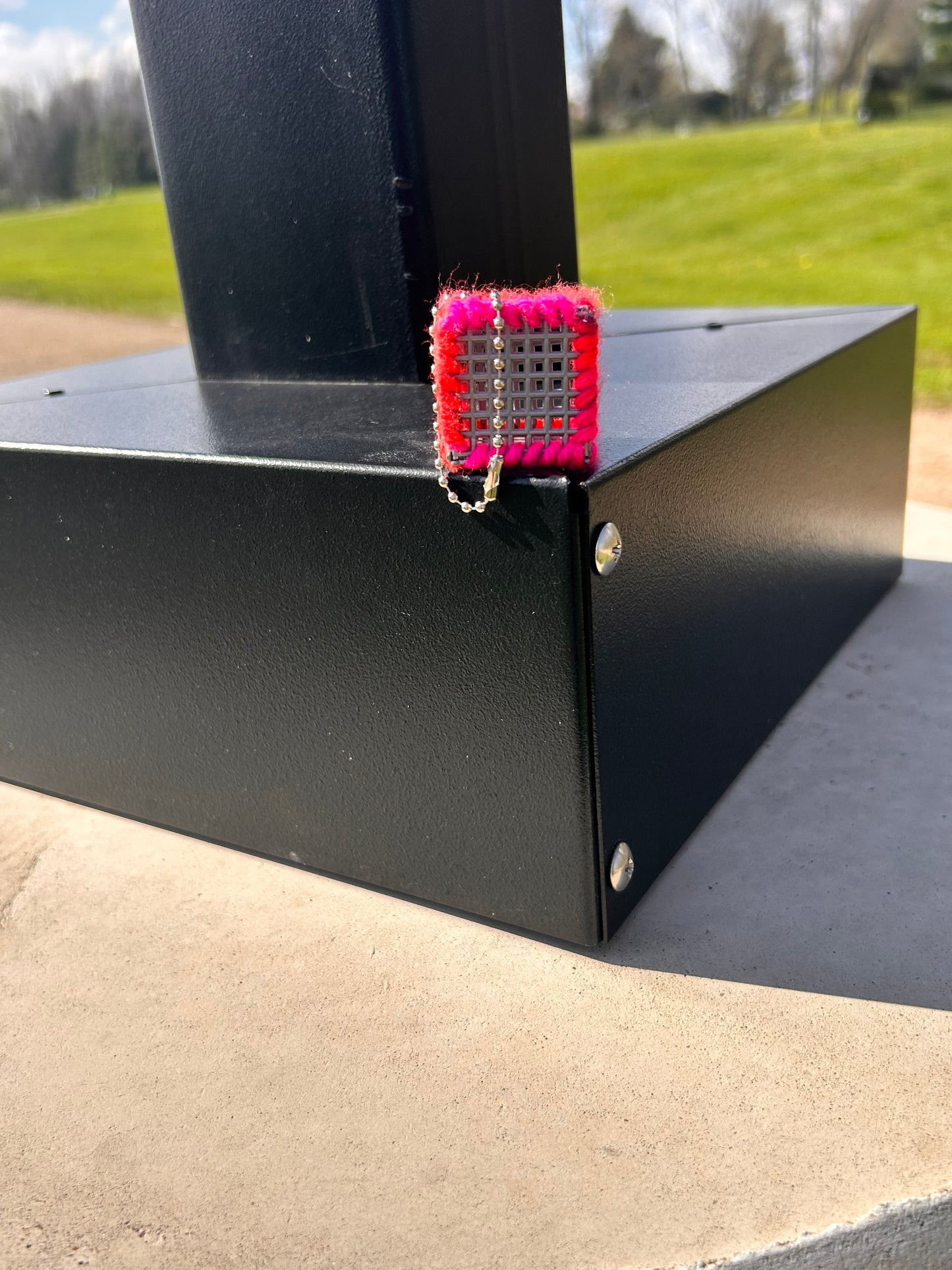 Handmade ASMR sensory cube laced with neon pink and orange yarn, 1.25 in. hexagon fidget art with ball chain clip pictured on the base of a lamp post at the park with lush green grass and trees in the background.