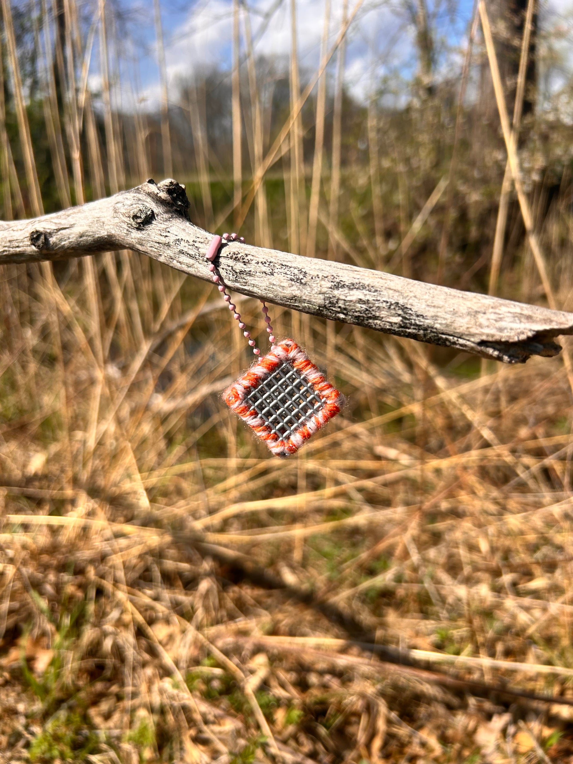 Handmade ASMR sensory cube laced with orange and cream yarn, 1.25 in. hexagon fidget art with ball chain clip hanging from a dead tree branch at the edge of the reeds by a stream.