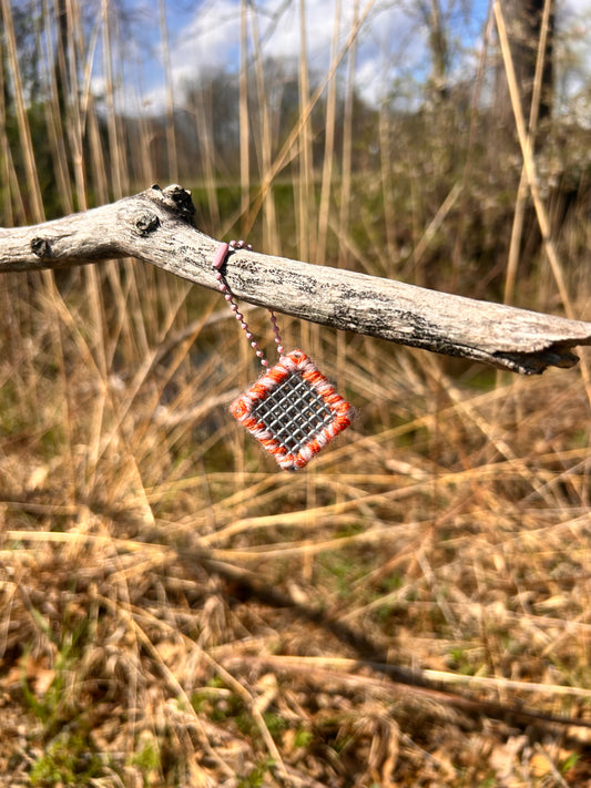 Handmade ASMR sensory cube laced with orange and cream yarn, 1.25 in. hexagon fidget art with ball chain clip hanging from a dead tree branch at the edge of the reeds by a stream.