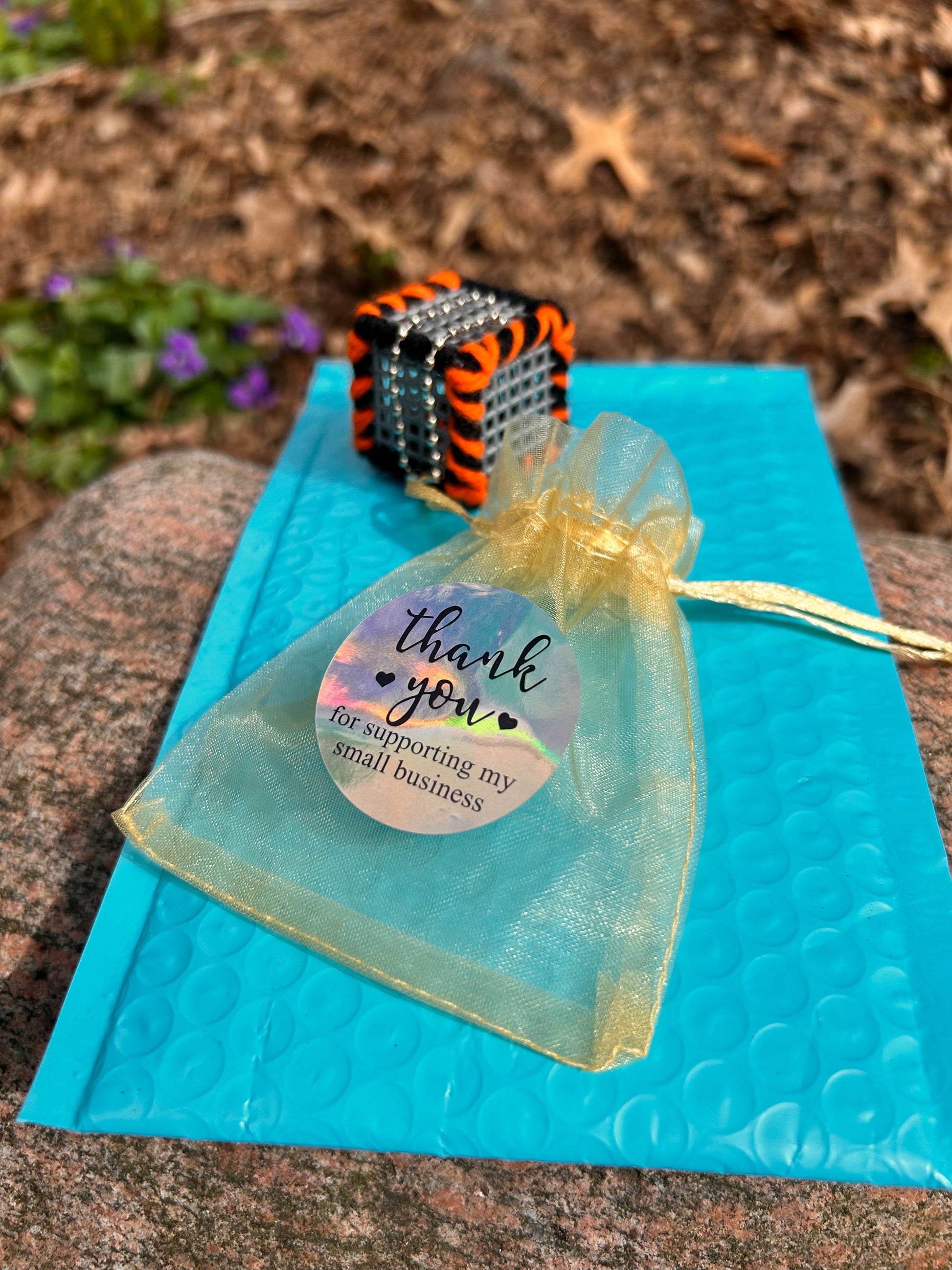 On top of a large rock in a nature setting there is a light blue bubble mailer with the Cincinnati Drive cube laced with orange and black yarn and a silver ball chain attached as well as a small gold mesh draw string gift bag with a thank you for supporting my small business sticker on the mesh bag.