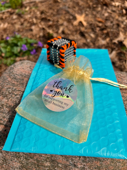 On top of a large rock in a nature setting there is a light blue bubble mailer with the Cincinnati Drive cube laced with orange and black yarn and a silver ball chain attached as well as a small gold mesh draw string gift bag with a thank you for supporting my small business sticker on the mesh bag.