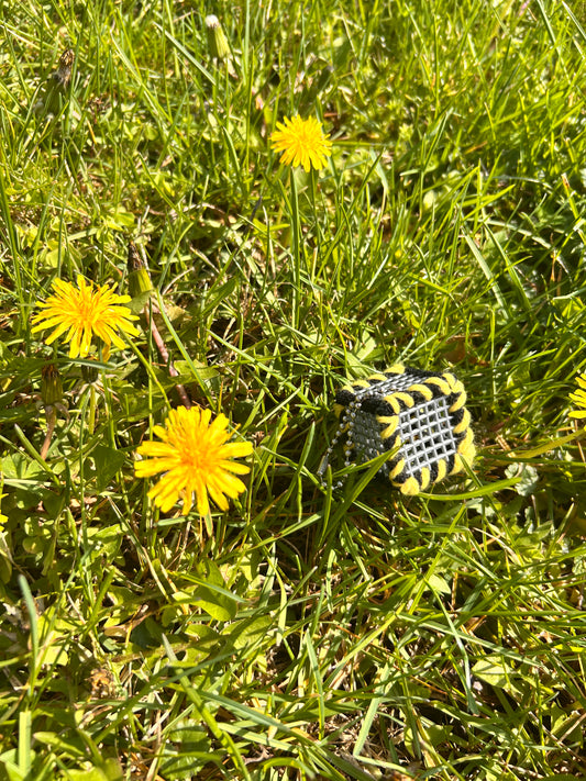 Handmade ASMR sensory cube laced with yellow and black yarn, 1.25 in. hexagon fidget art with ball chain clip pictured in the grass next to 3 dandelion blooms.