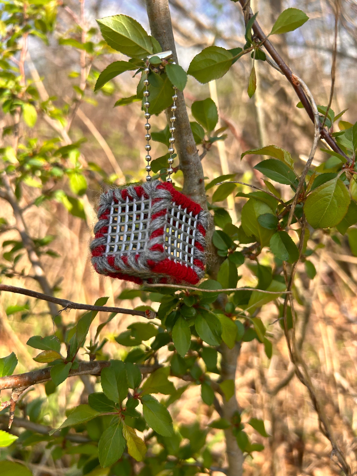Handmade ASMR sensory cube laced with Scarlet and Gray yarn, 1.25 in. hexagon fidget art with ball chain clip pictured on a tree branch with lots of small leaves around the cube in a sun filtered wooded area.
