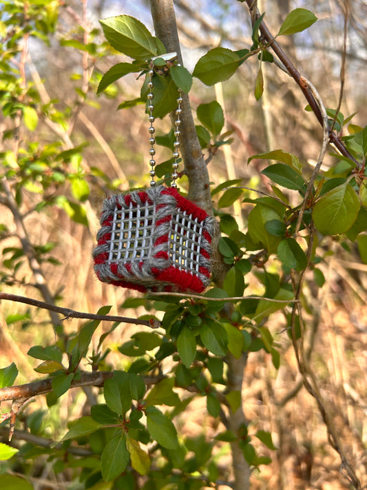Handmade ASMR sensory cube laced with Scarlet and Gray yarn, 1.25 in. hexagon fidget art with ball chain clip pictured on a tree branch with lots of small leaves around the cube in a sun filtered wooded area.