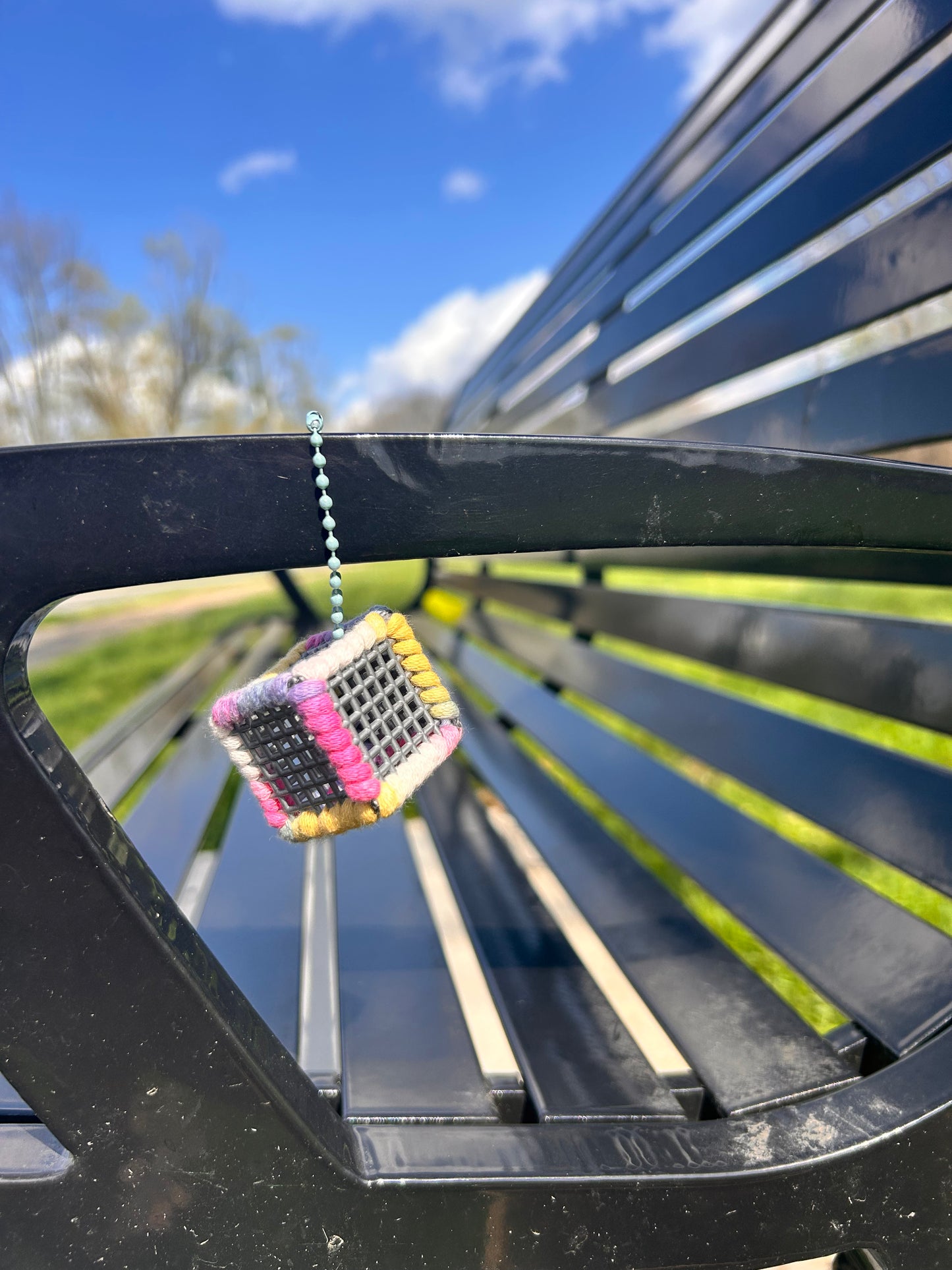 Handmade ASMR sensory cube laced with variegated pastel colored yarn, 1.25 in. hexagon fidget art with ball chain clip pictured hanging on the arm rest of a black metal bench with a blue sky, white clouds, green grass and trees in the background on a sunny day.