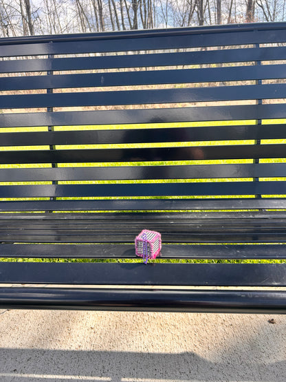 Handmade Pink and purple yarn laced Flex-Hex-Sensi © key chain - AMSR / Sensory cube with a purple ball chain sitting on a black metal bench in the sun with grass and trees in the background.
