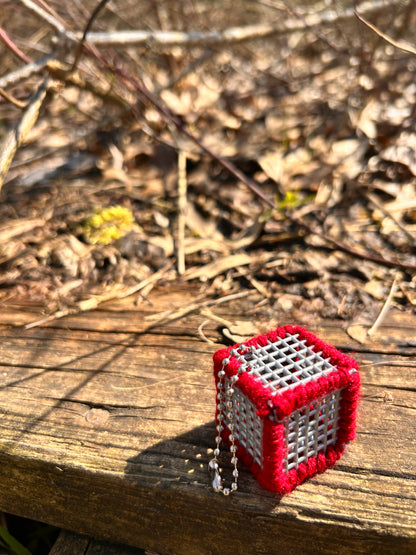Handmade small red and silver 1.25 inch ASMR / Sensory cube-shaped object on a wooden beam at the park with a forest floor background.