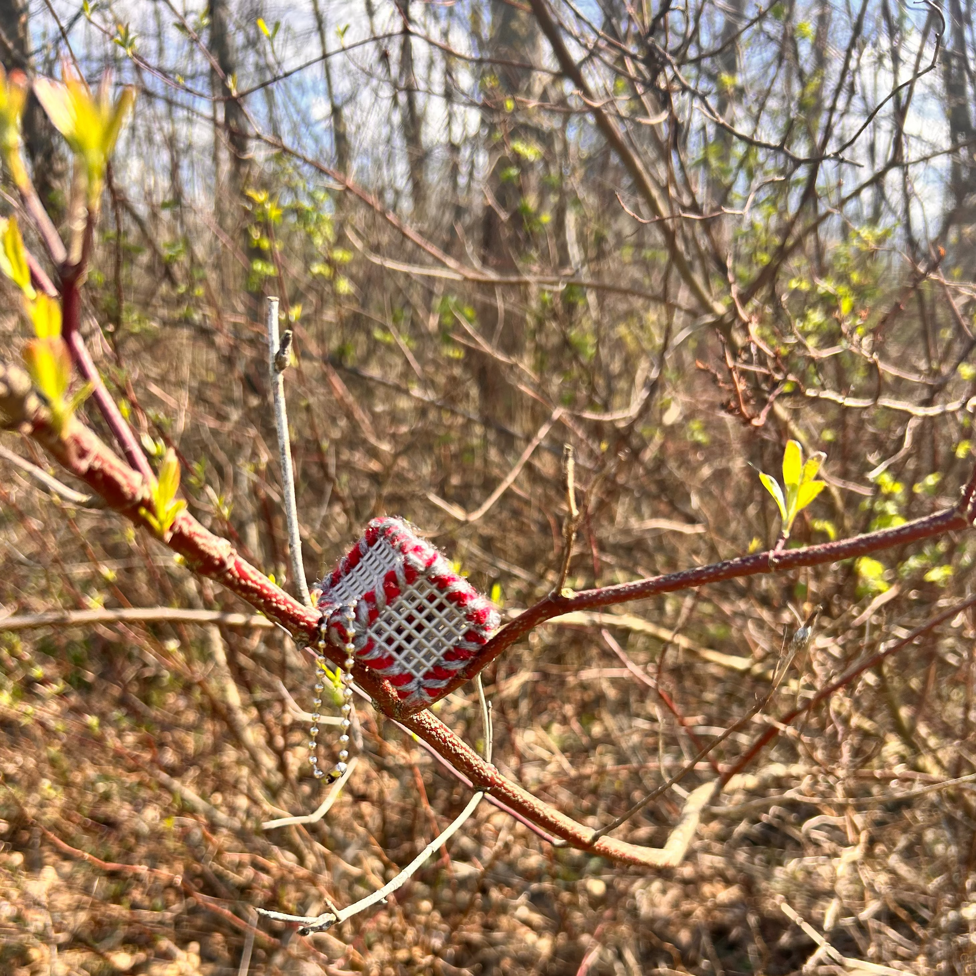 Handmade ASMR sensory cube laced with Scarlet and Gray yarn, 1.25 in. hexagon fidget art with ball chain clip pictured on a tree branch in a wooded area.
