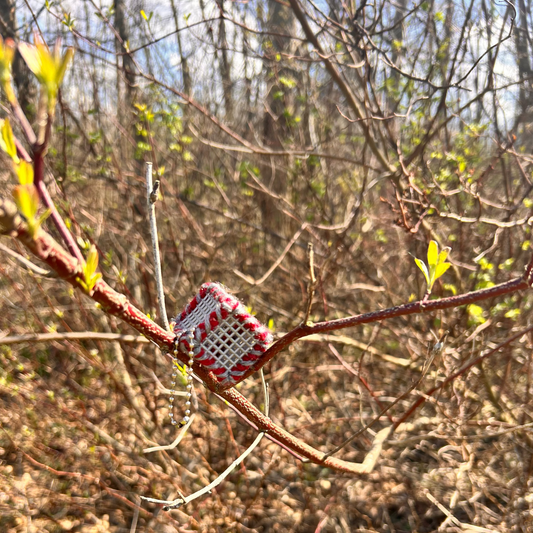 Handmade ASMR sensory cube laced with Scarlet and Gray yarn, 1.25 in. hexagon fidget art with ball chain clip pictured on a tree branch in a wooded area.