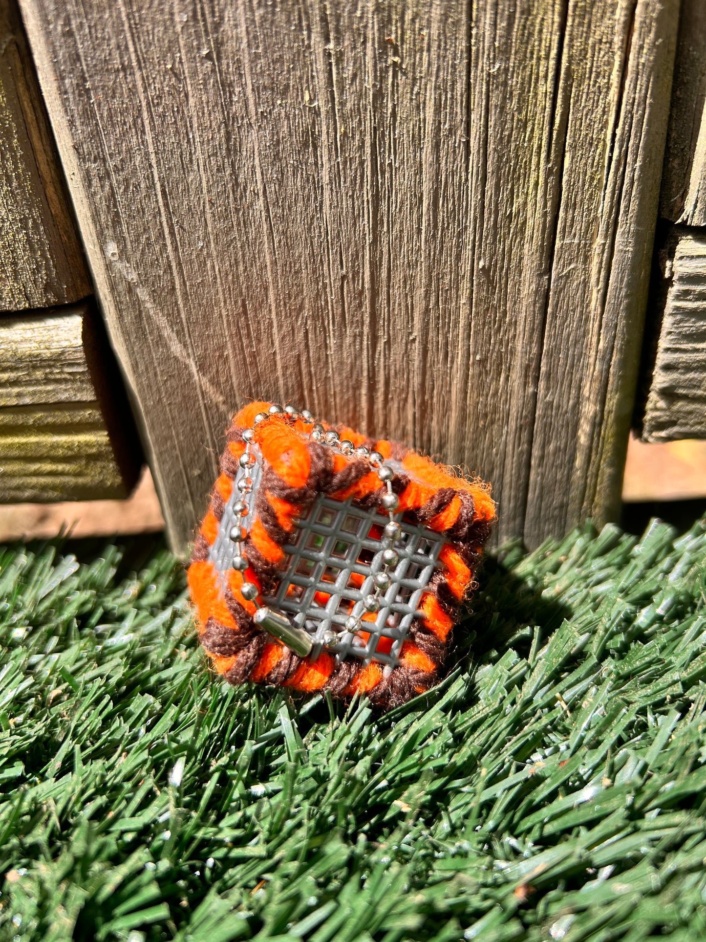 Handmade ASMR sensory art cube measuring 1.25 inches laced with orange and brown yarn with an attached silver ball chain sitting in the sun on artificial turf.