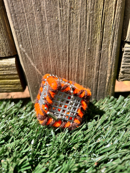 Handmade ASMR sensory art cube measuring 1.25 inches laced with orange and brown yarn with an attached silver ball chain sitting in the sun on artificial turf.