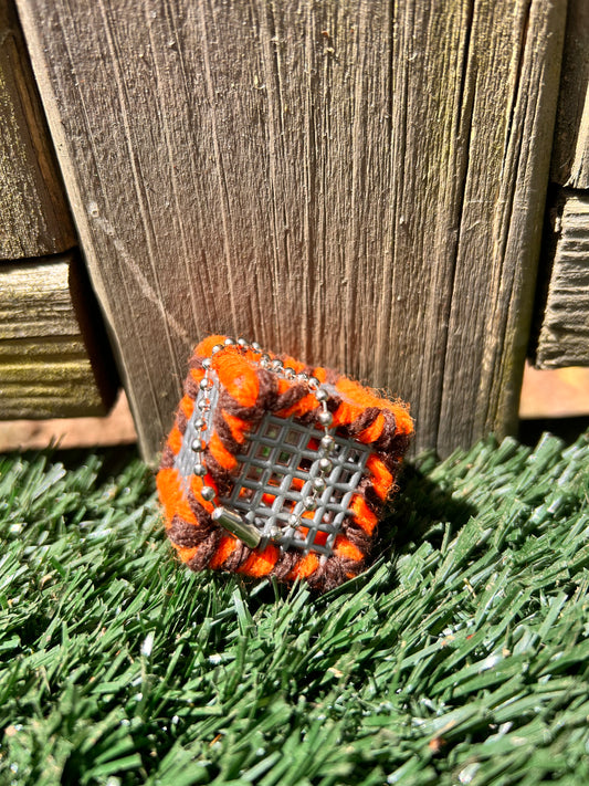 Handmade ASMR sensory art cube measuring 1.25 inches laced with orange and brown yarn with an attached silver ball chain sitting in the sun on artificial turf.