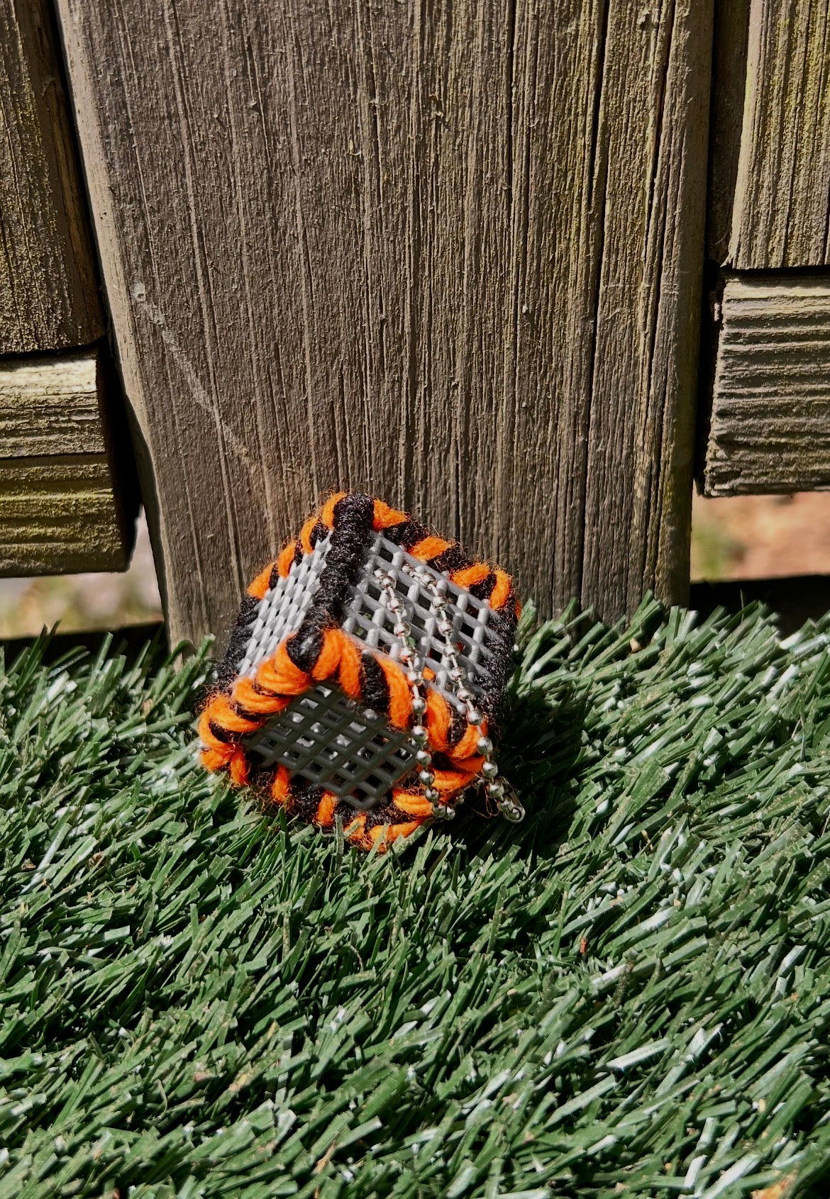 Handmade ASMR sensory cube laced with black and orange yarn, 1.25 in. hexagon fidget art with ball chain clip pictured sitting outside in the sun on artificial turf.