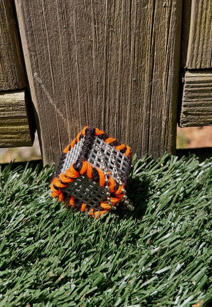 Handmade ASMR sensory cube laced with black and orange yarn, 1.25 in. hexagon fidget art with ball chain clip pictured sitting outside in the sun on artificial turf.